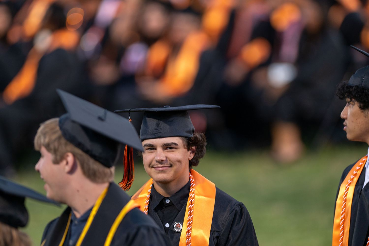 Graduates in black caps and golden gowns stand in a row during an outdoor commencement ceremony.