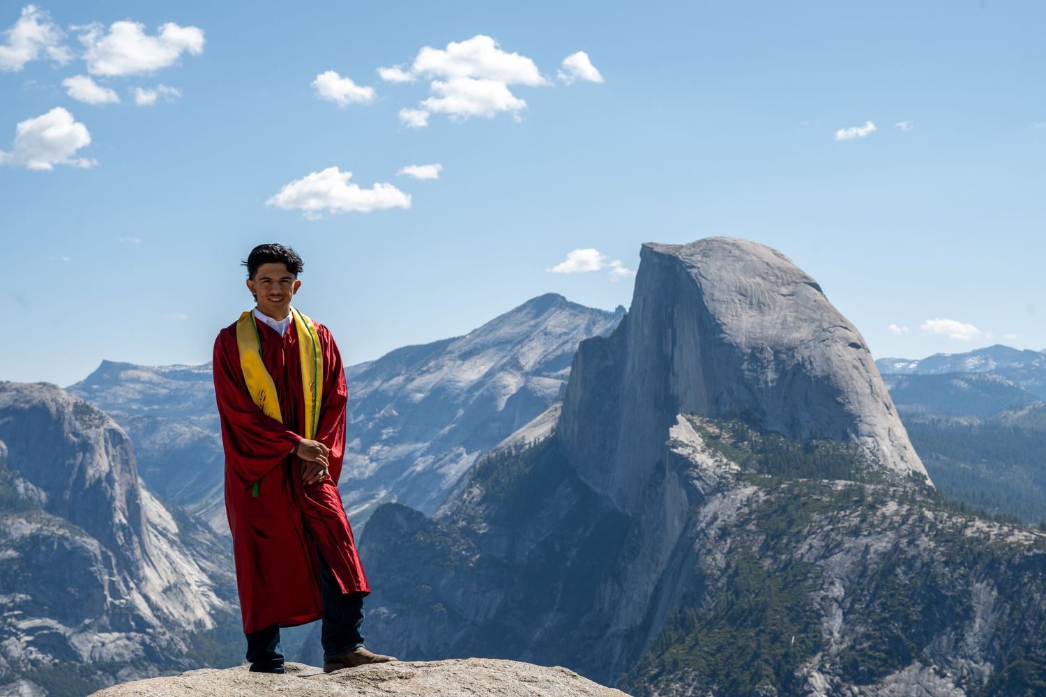 A figure in red outerwear stands atop a mountain peak overlooking the stunning Half Dome rock formation in Yosemite.