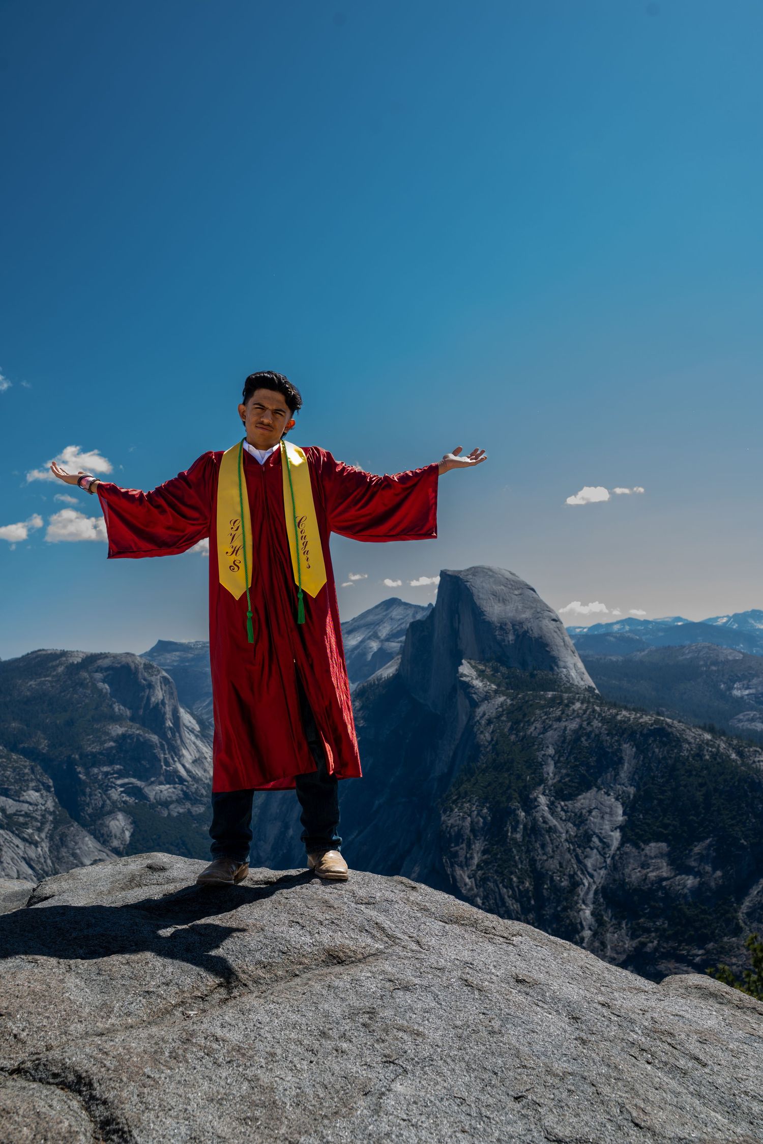A person in graduation robes stands on Glacier Point with Half Dome and Yosemite Valley's granite peaks in the background.