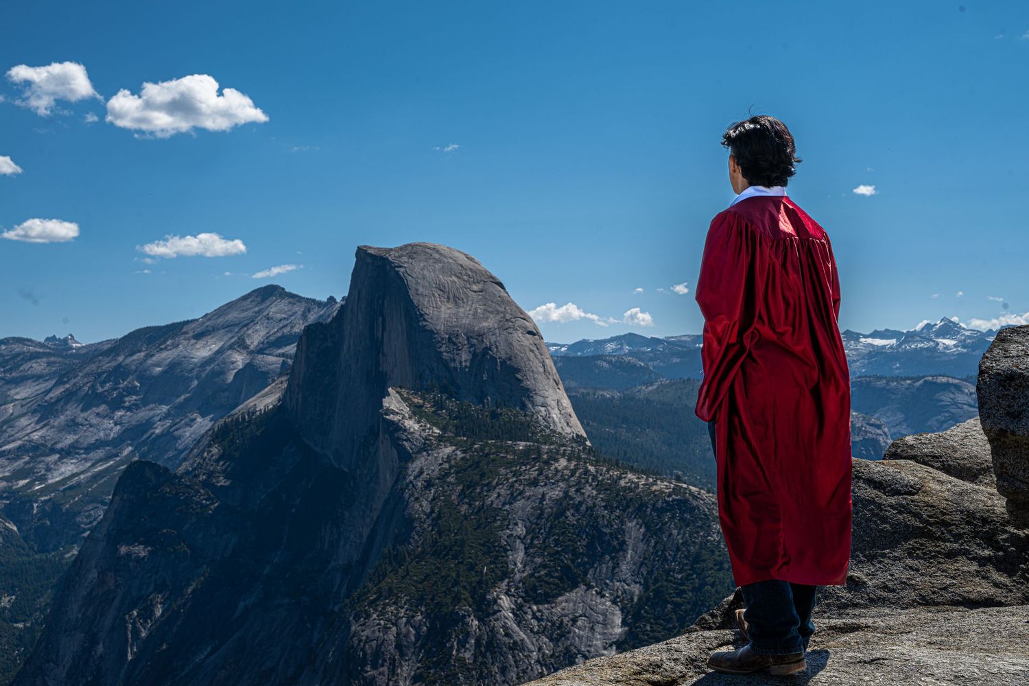 A silhouetted person in bright red jacket stands on granite peak with Half Dome and Yosemite Valley vista in background.
