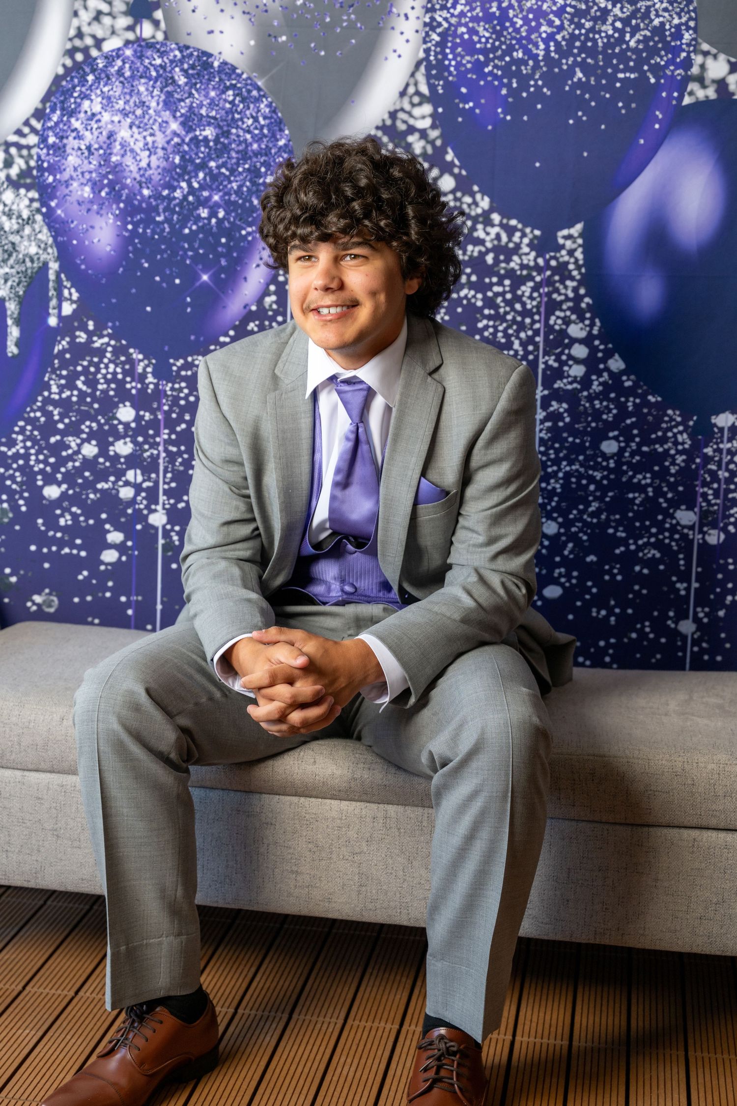 Formal portrait against blue and silver balloon backdrop showing grey suit with purple tie and vest for special occasion.