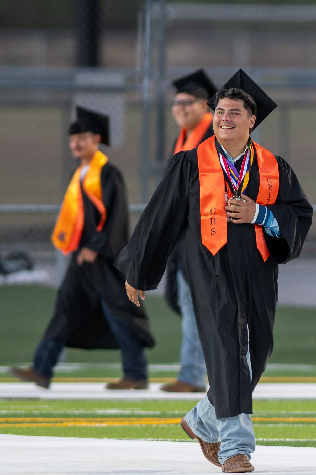 Smiling graduate in black gown and orange stole walks across football field during outdoor graduation ceremony.