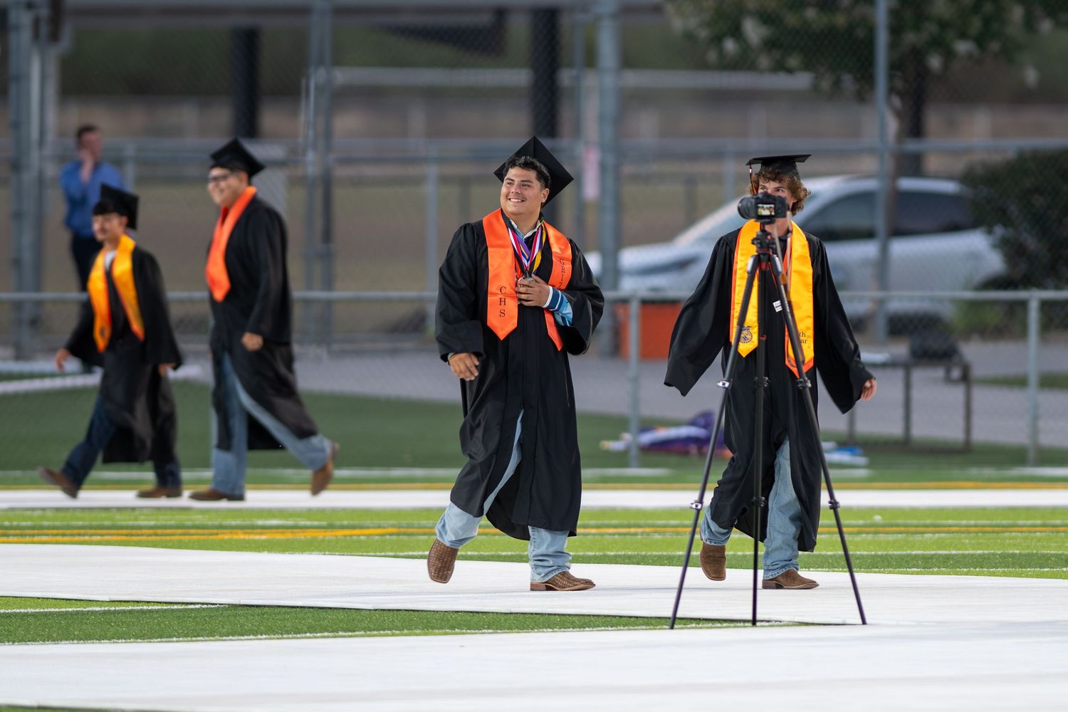 Graduates in black robes and orange sashes walk across a field during an outdoor graduation ceremony.