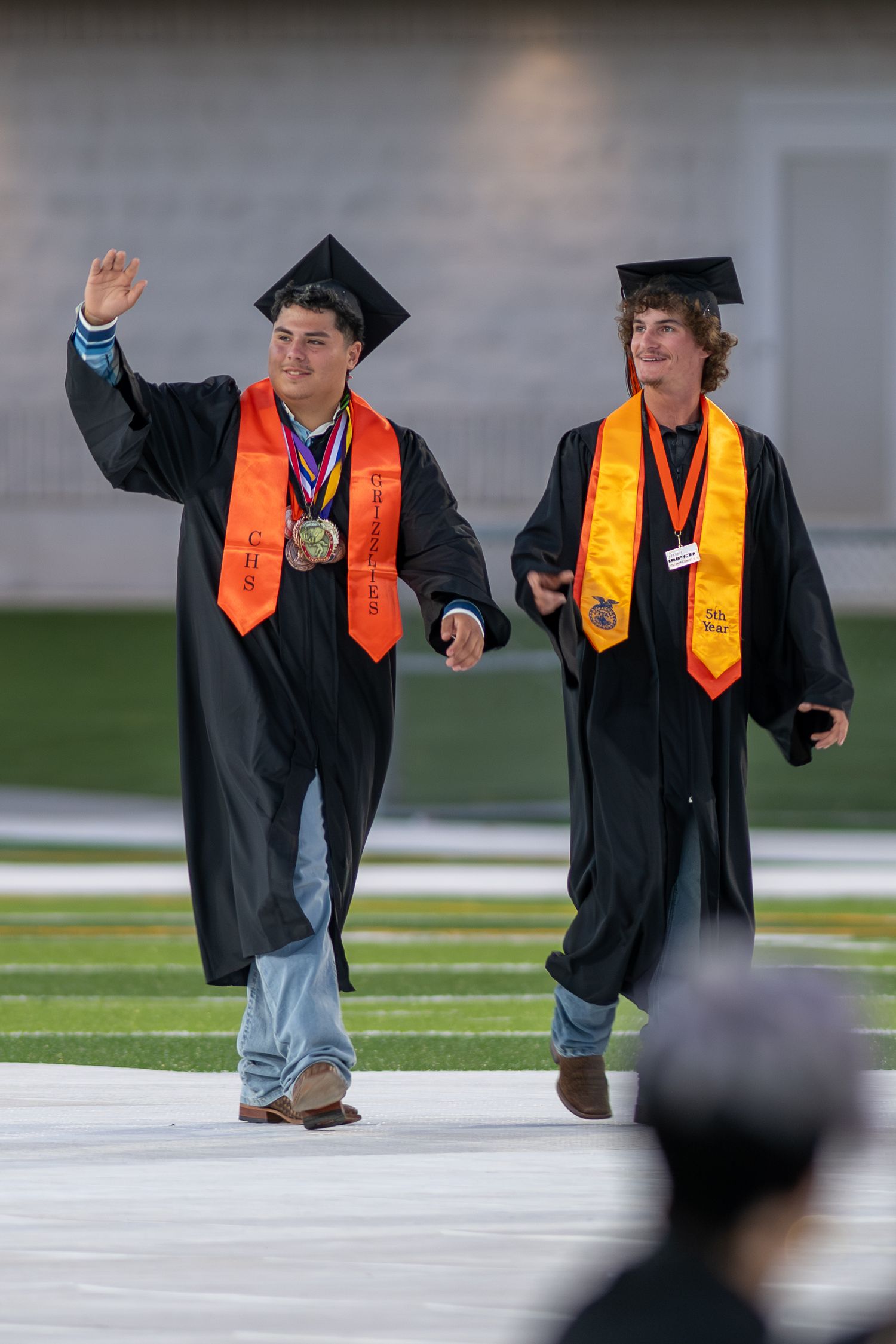 Two graduates in black robes and orange stoles walk across a football field during a graduation ceremony.