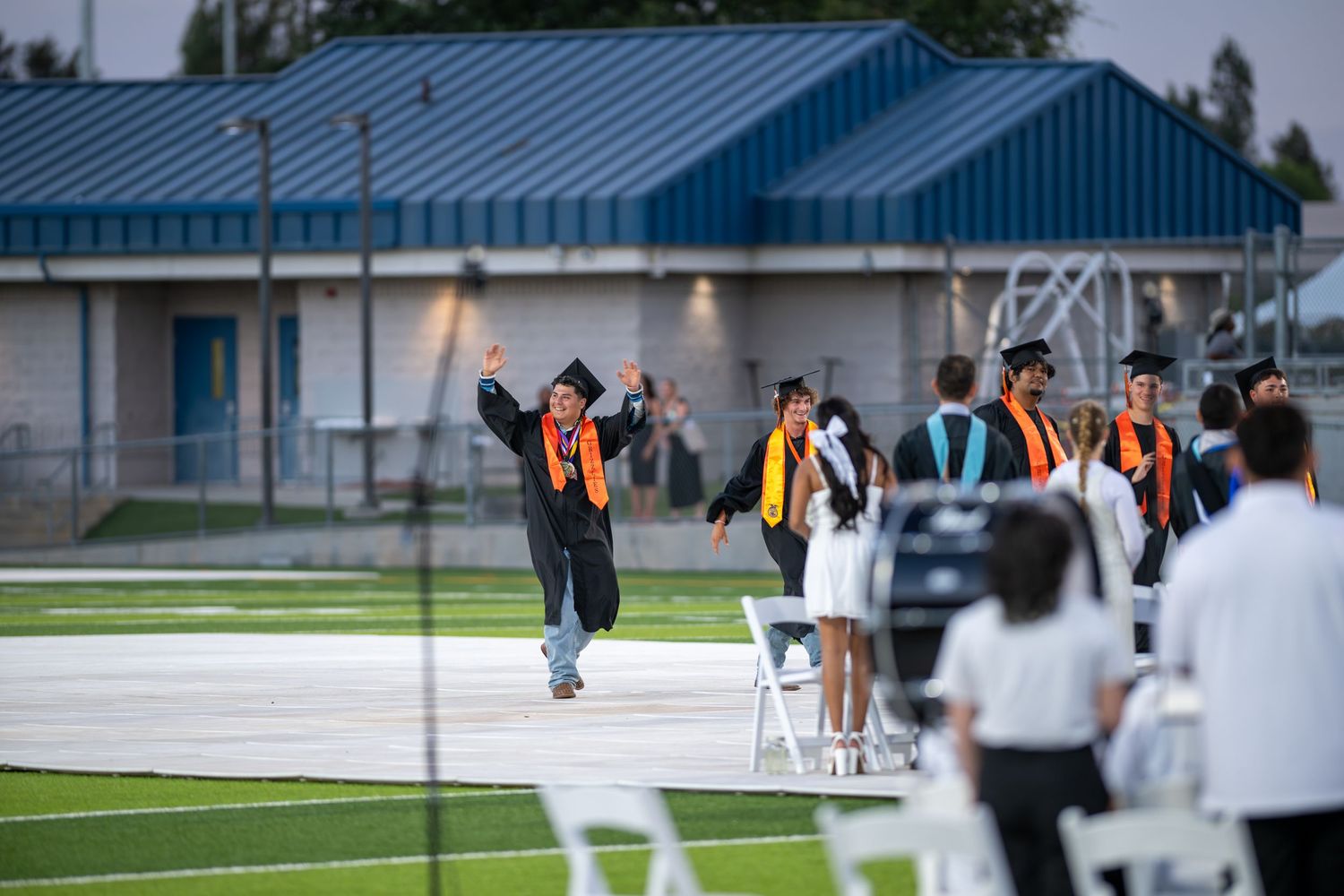 High school graduates in black and orange caps and gowns walk during an outdoor graduation ceremony on a green field.