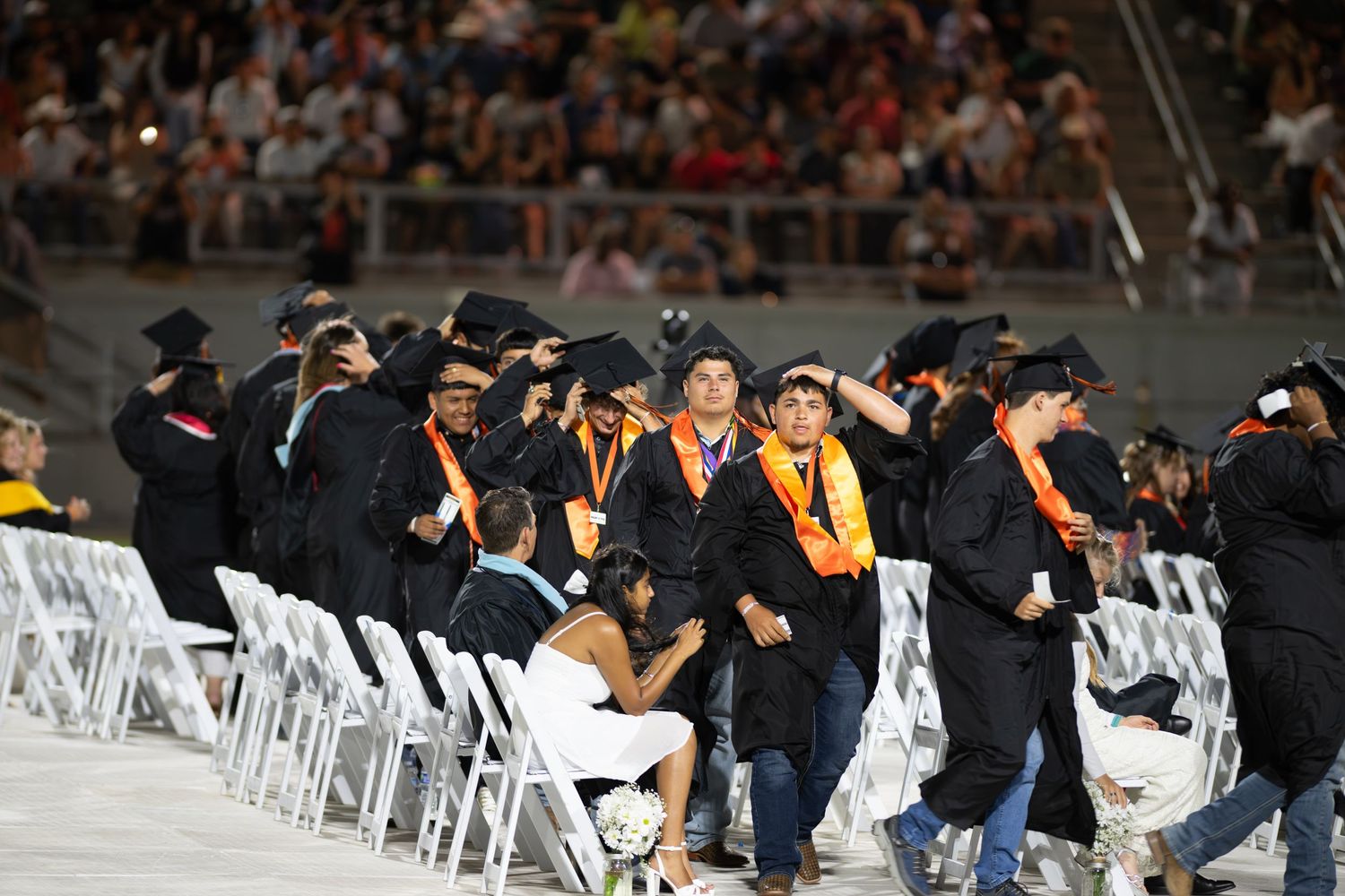 Graduates in black caps and gowns with orange sashes walk in procession at an outdoor graduation ceremony.