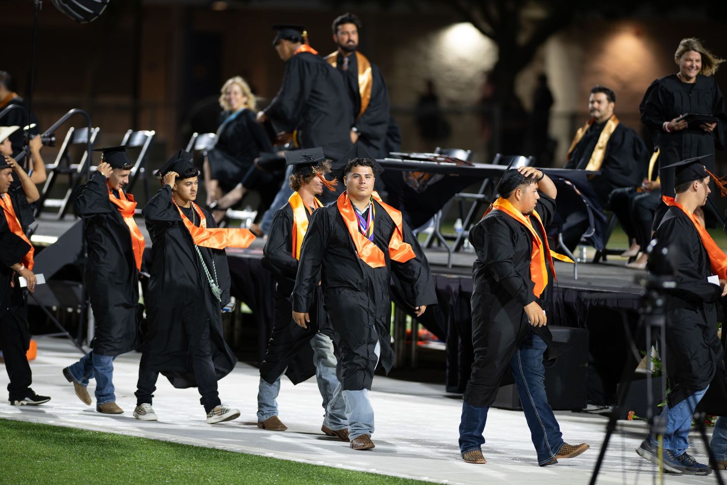 Graduates in black and orange robes walk in procession at an outdoor graduation ceremony on a football field at night.
