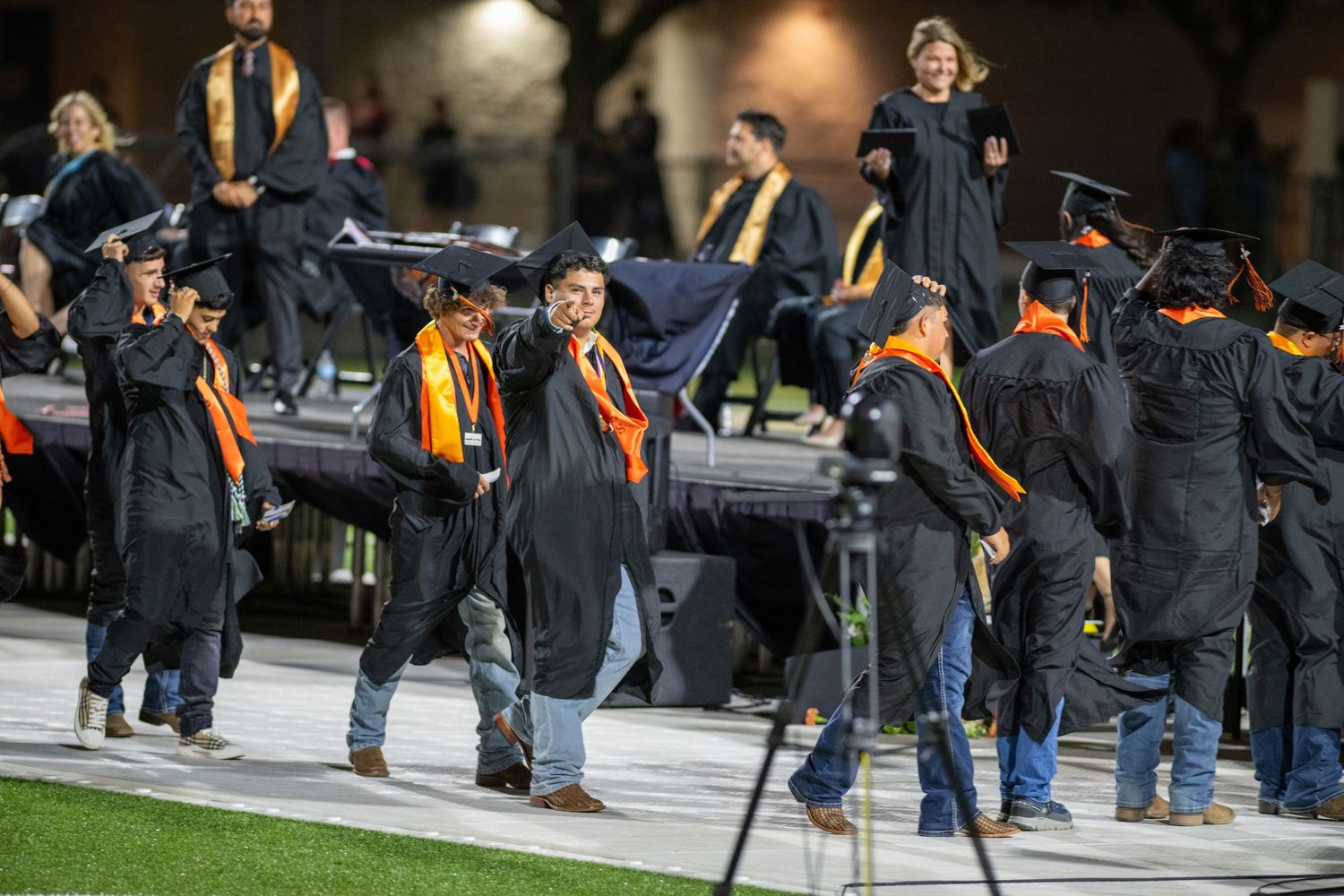 Graduates in black and orange robes celebrate at an outdoor graduation ceremony on a stadium field at night.