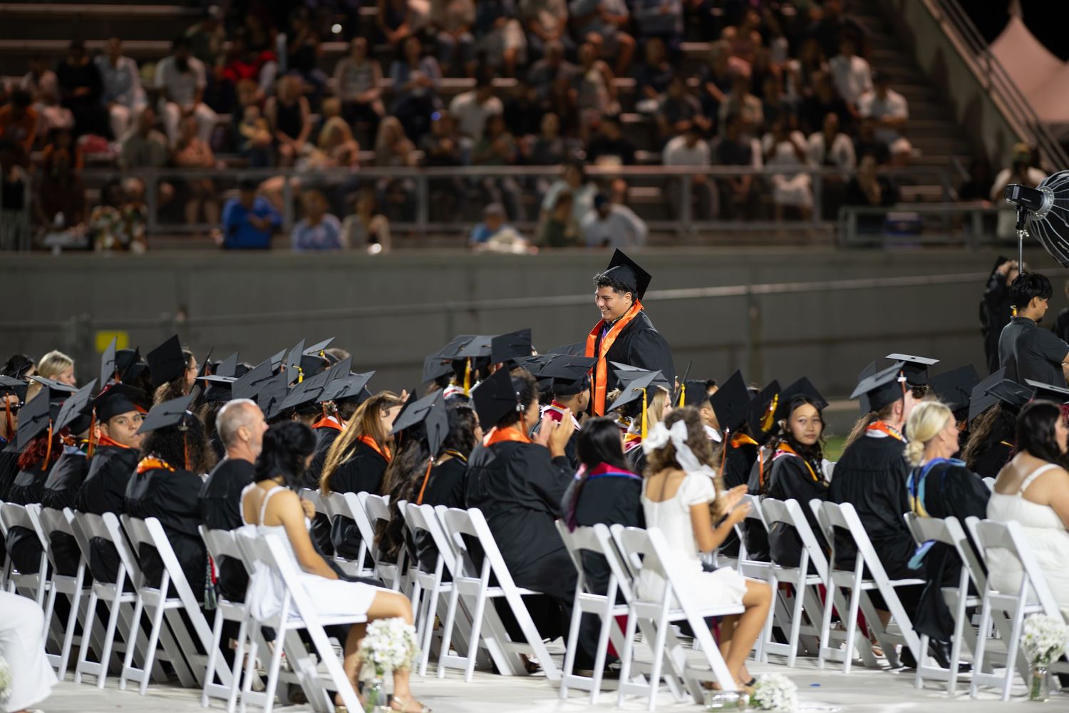 Students in graduation caps and gowns seated at an indoor commencement ceremony with audience in background.