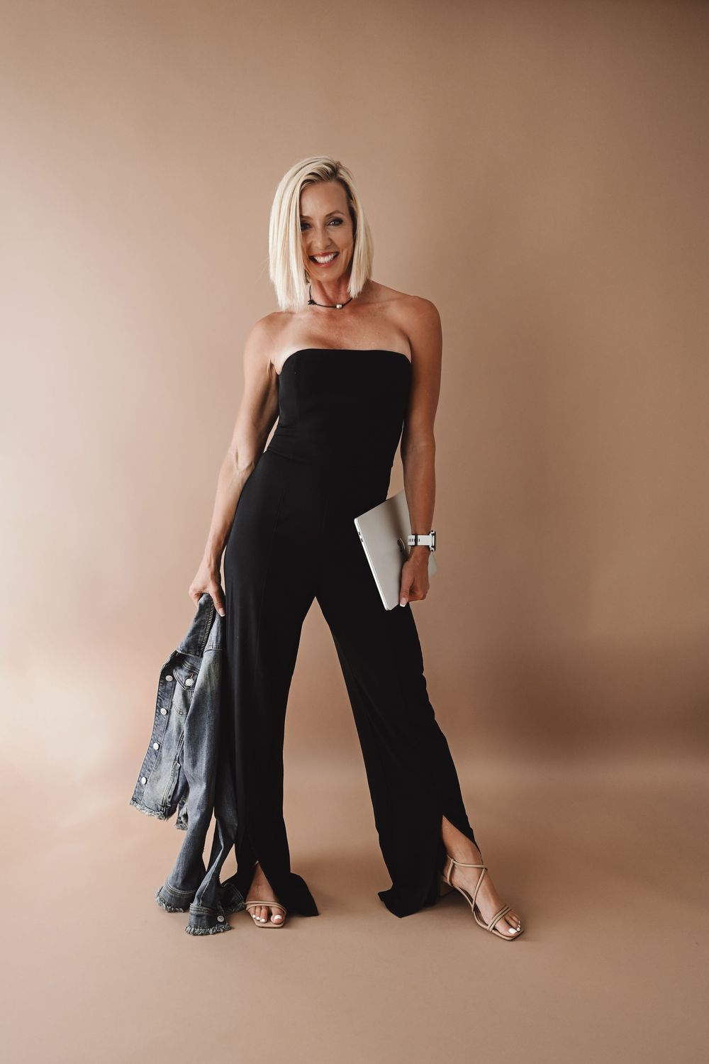 Woman in black jumpsuit poses against beige backdrop with denim jacket.