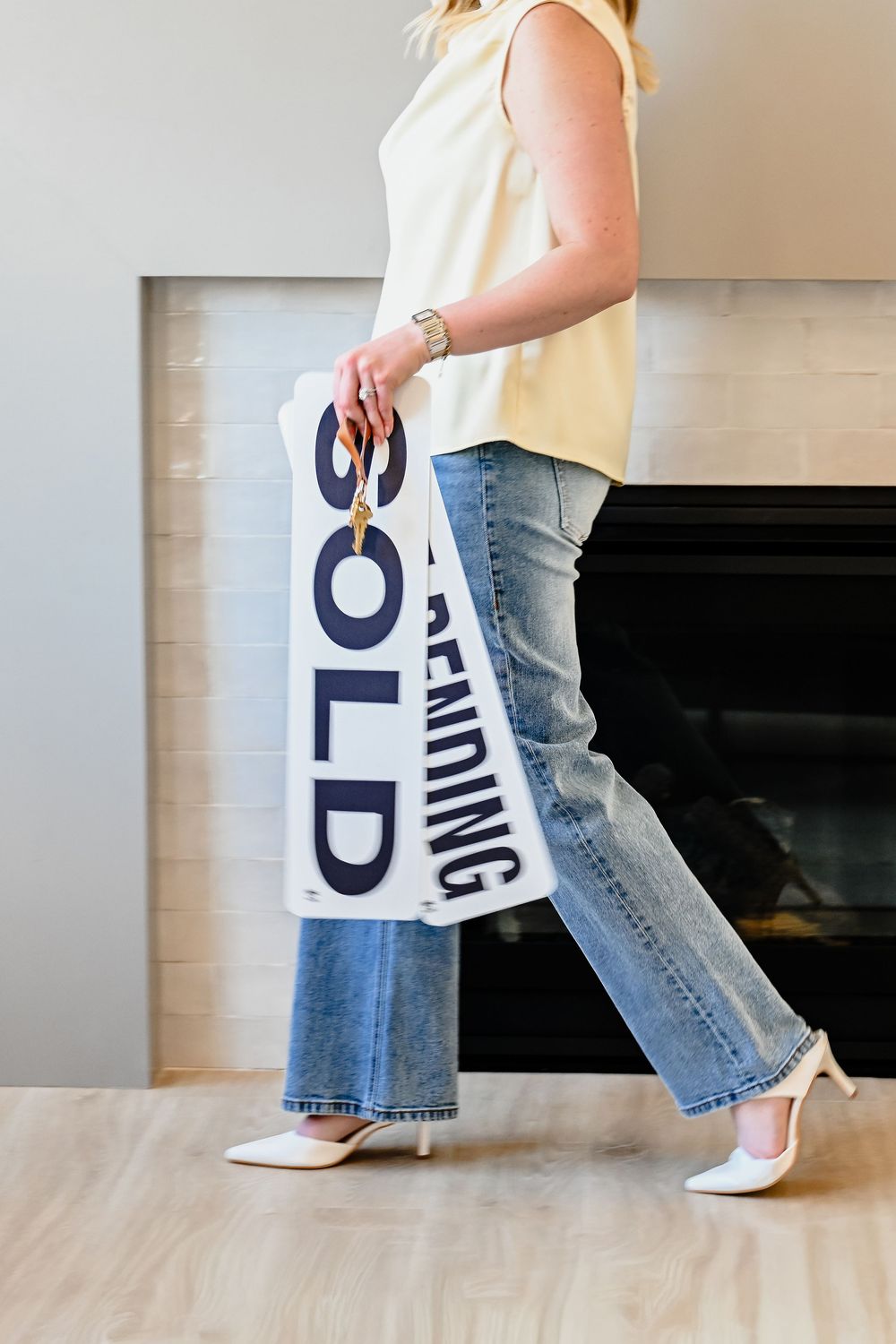 A woman in jeans and heels carries a SOLD and PENDING real estate sign inside a home with a fireplace.
