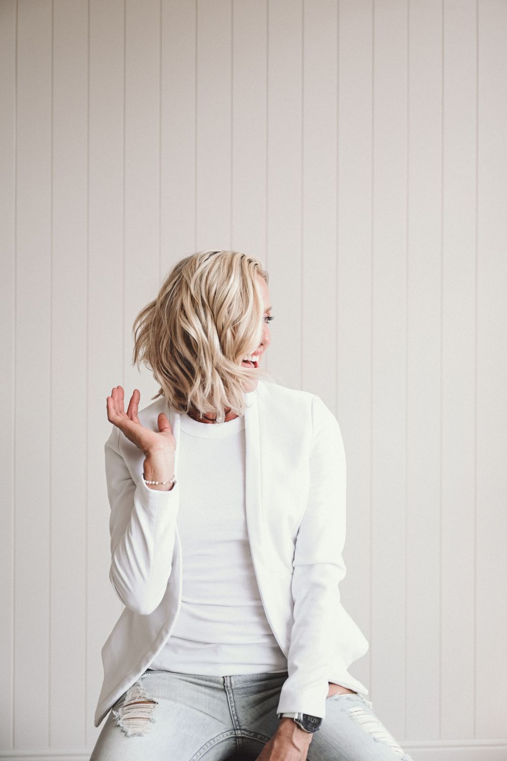 Person in a white blazer poses candidly against a white wall in a minimalist studio setting.