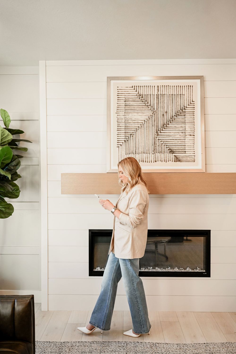 Woman in beige blazer and flared jeans walking past a modern fireplace with geometric wall art above.