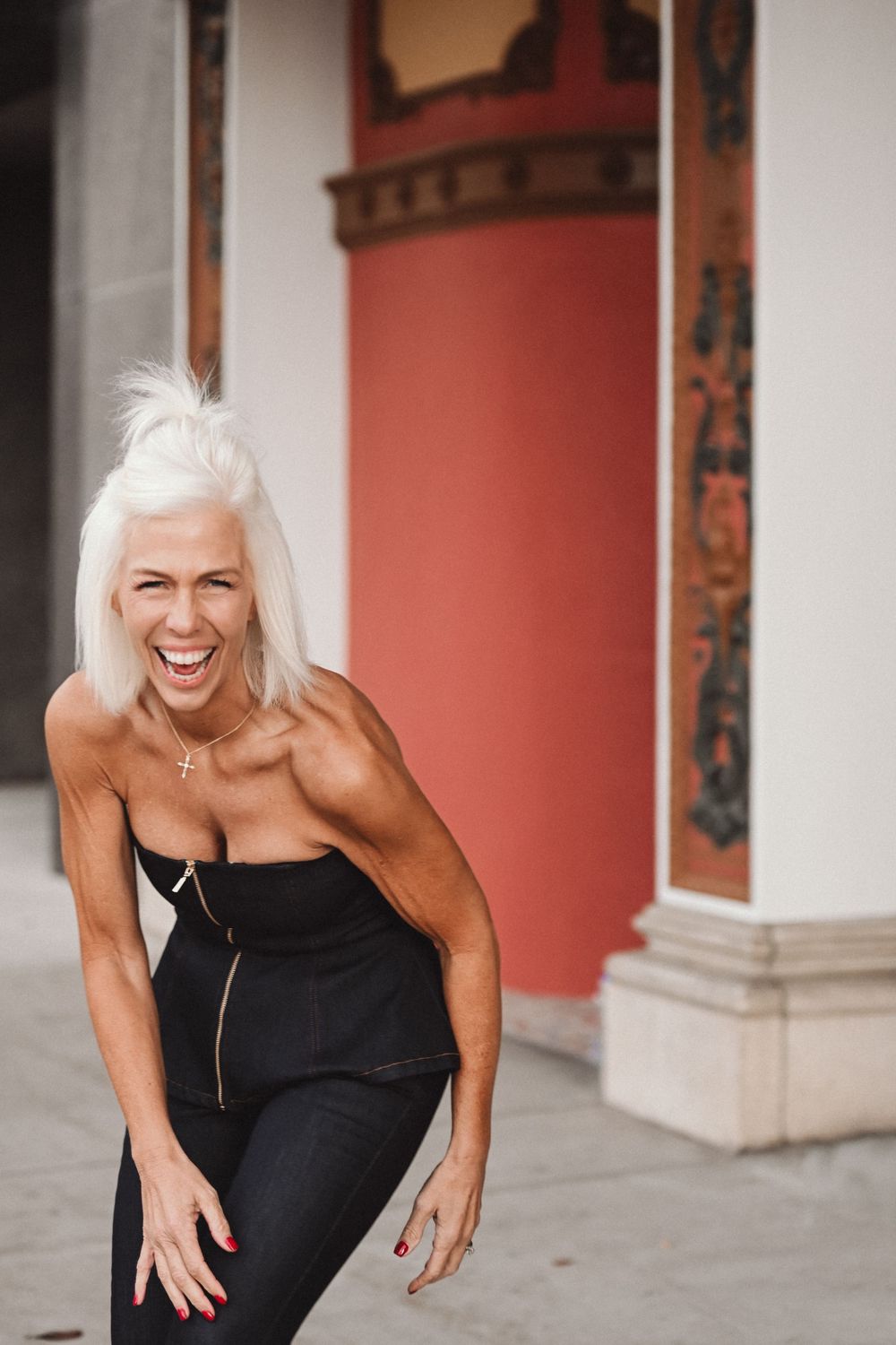 Woman with white hair wearing black dress laughs joyfully while posing against red and white building exterior.