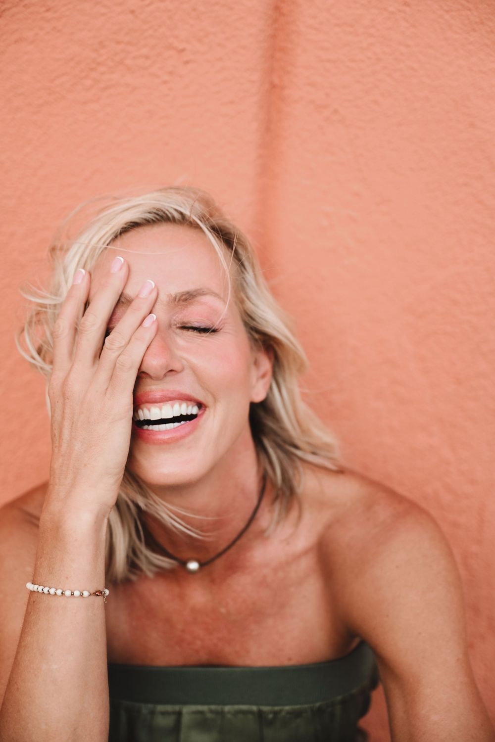 Woman laughing with hand covering face against coral wall wearing pearl necklace.