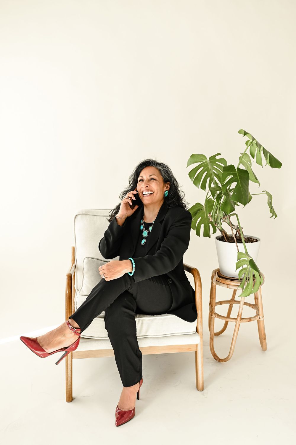 Woman in black suit and red heels sits in armchair, smiling while talking on phone, with monstera plant nearby.