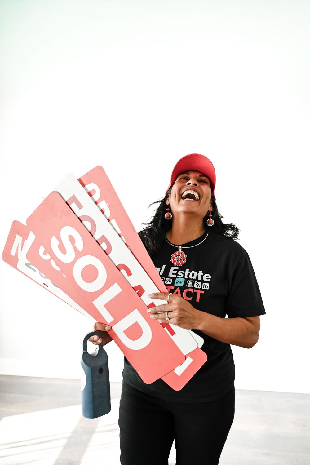 Excited real estate agent laughing while holding multiple red SOLD signs, wearing a black t-shirt and red cap.