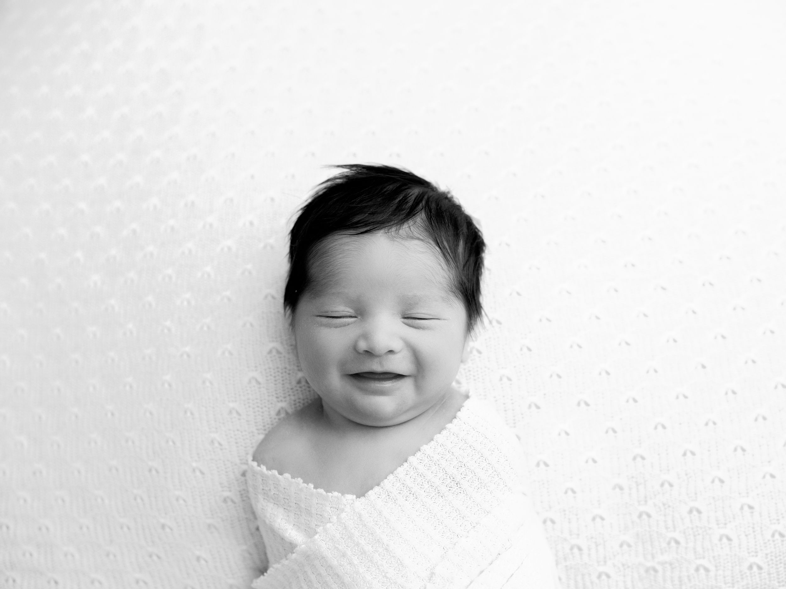 Black and white portrait of a swaddled newborn baby with a joyful smile on a white textured background.