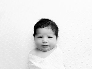 A small baby with chubby cheeks and a content expression peers at the camera on a white background.