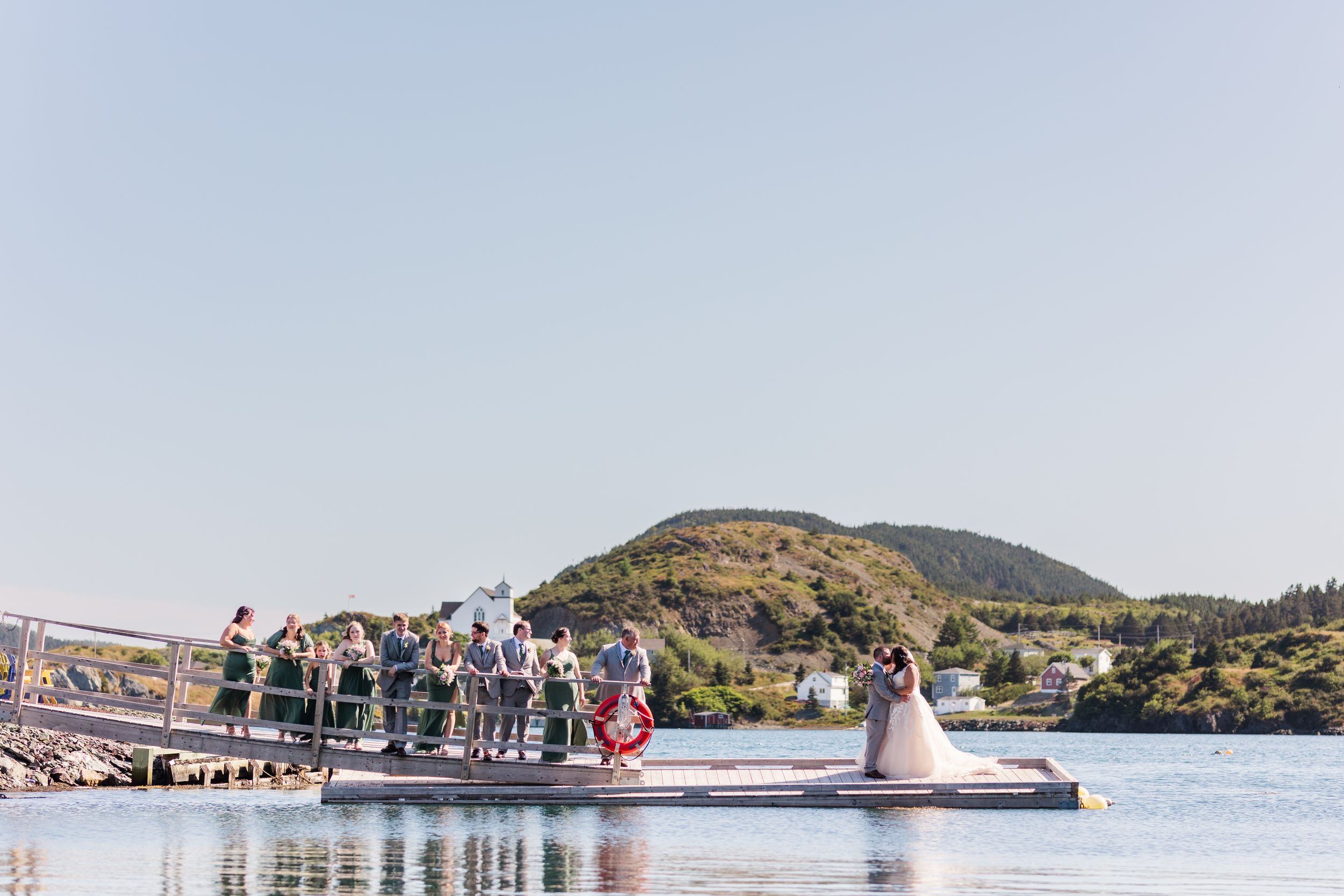 Wedding party gathers on a wooden dock over a scenic lake with mountains in the background on a clear day.