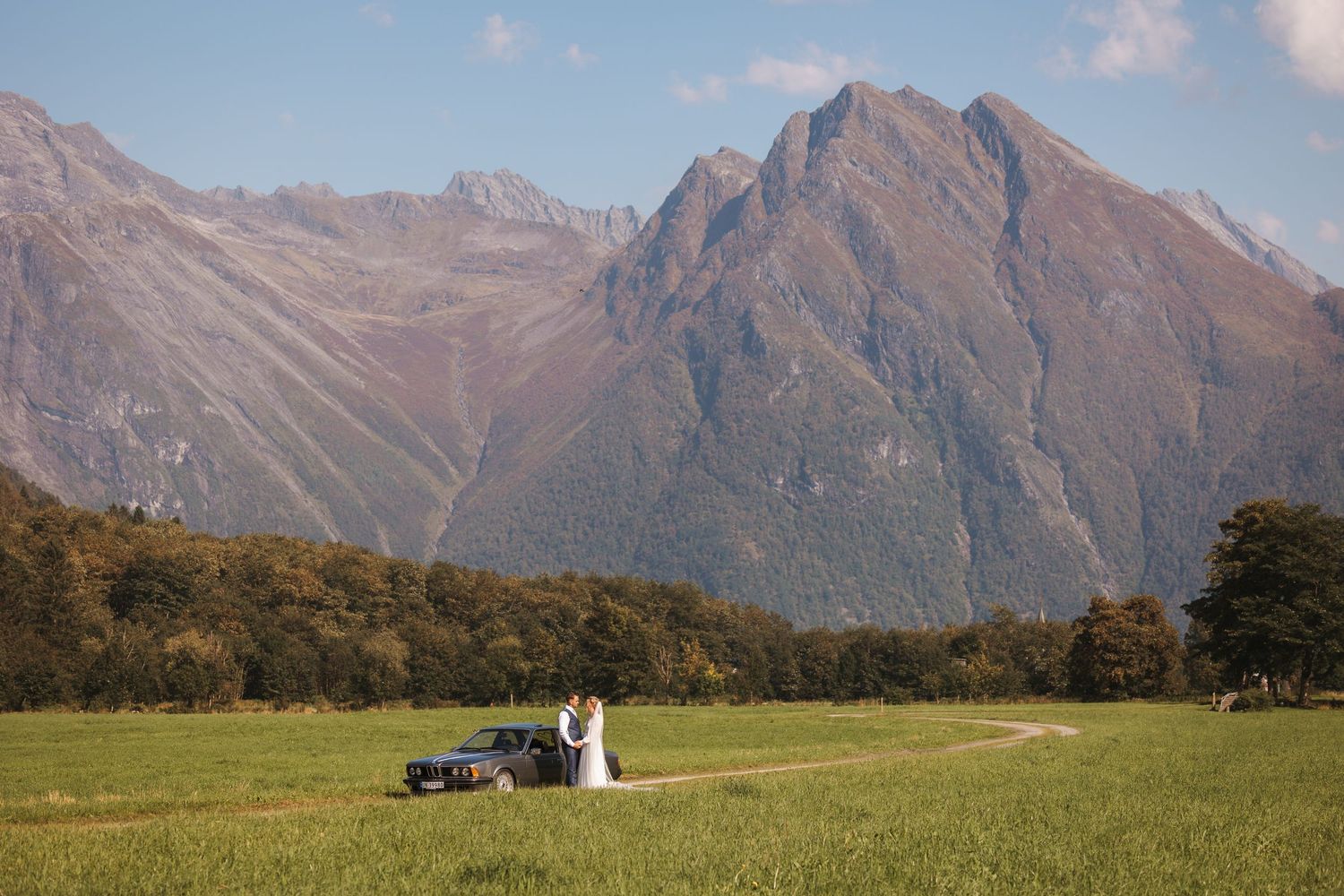A couple sits on blanket in grassy field with majestic mountain peaks rising in the background under soft clouds.