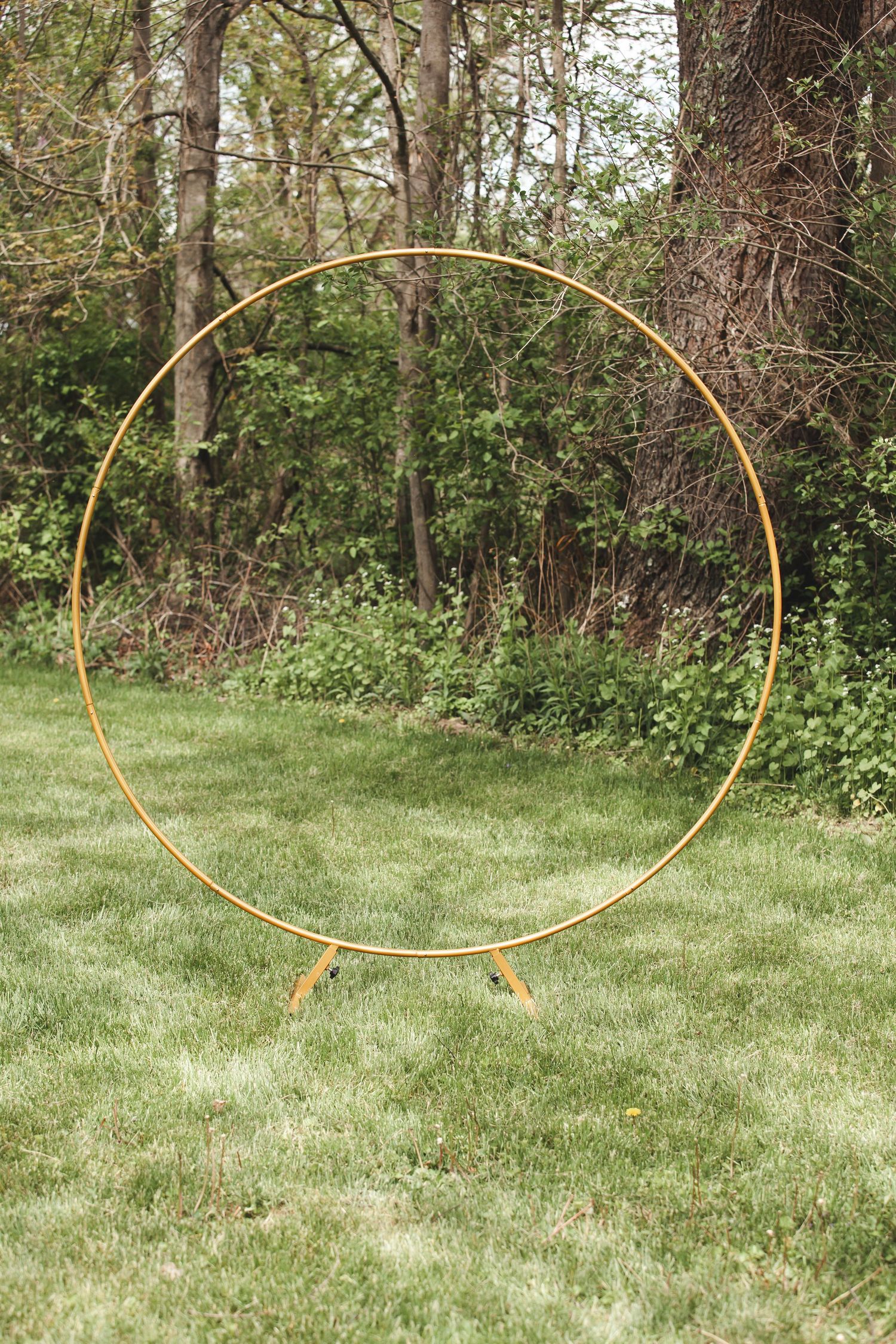Large gold circular wedding arch or hoop stands on grass lawn against wooded backdrop in outdoor garden setting.