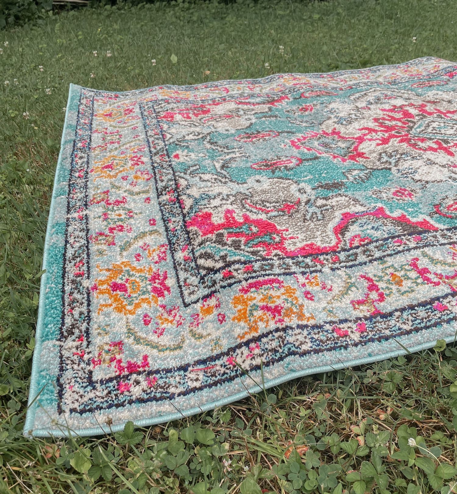 A vintage-style area rug with faded turquoise, pink and yellow patterns lies on green grass outdoors.