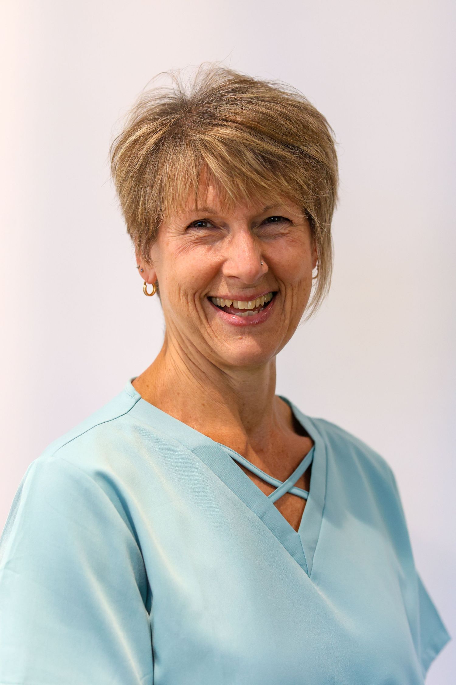 Healthcare professional wearing light blue scrubs smiles warmly against neutral background.