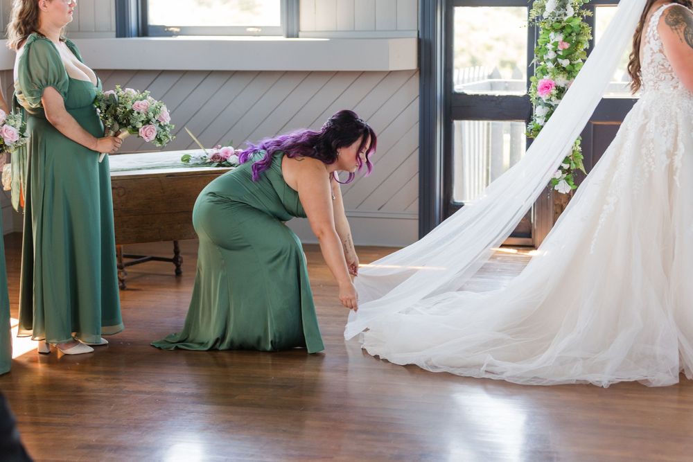 Bridesmaids in sage green dresses adjust a long white wedding veil on a hardwood floor.