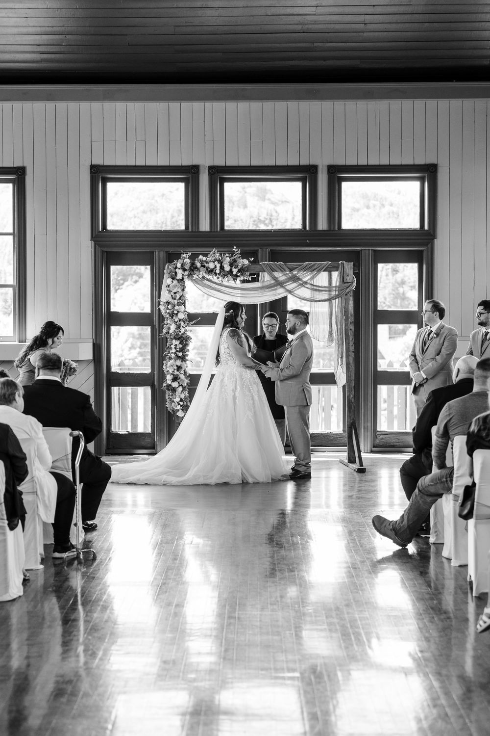 Indoor wedding ceremony captured in black and white with an elegant floral arch and polished hardwood floors.