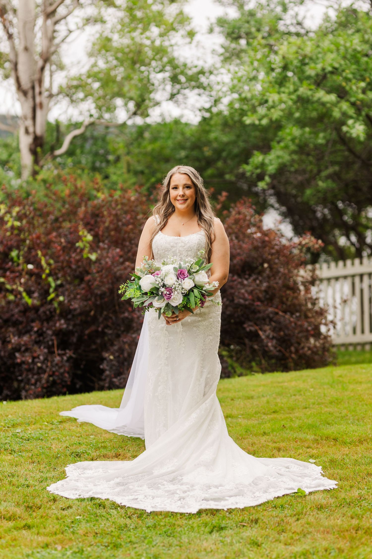 A bride in a stunning white lace gown with a flowing train holds a blush and white bouquet in a garden setting.