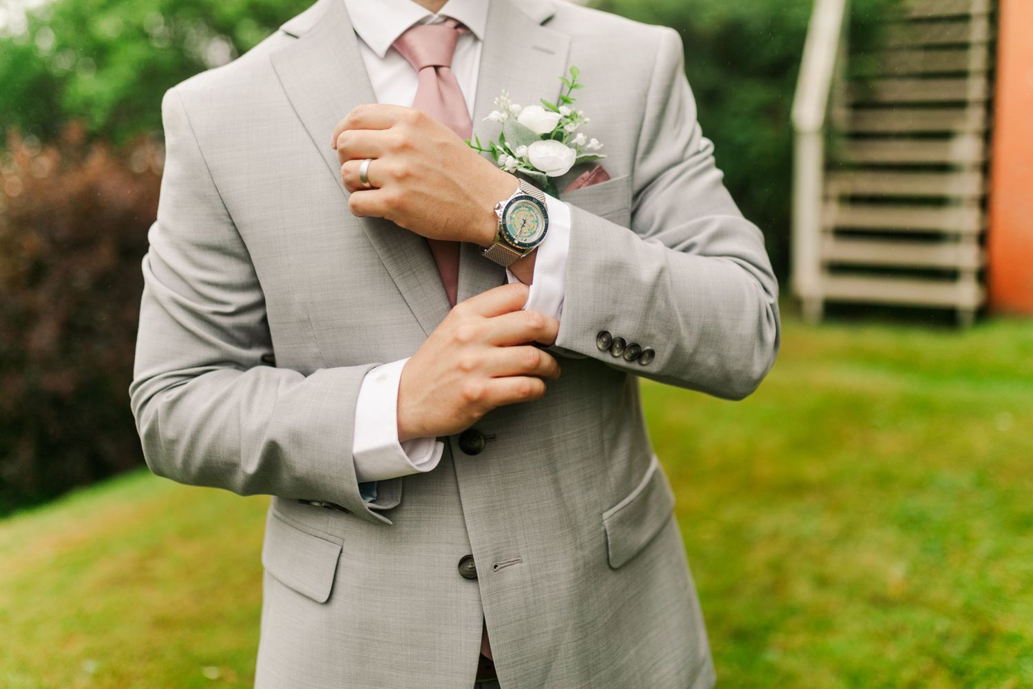 A person wearing a light gray suit adjusts their tie against a green outdoor background.