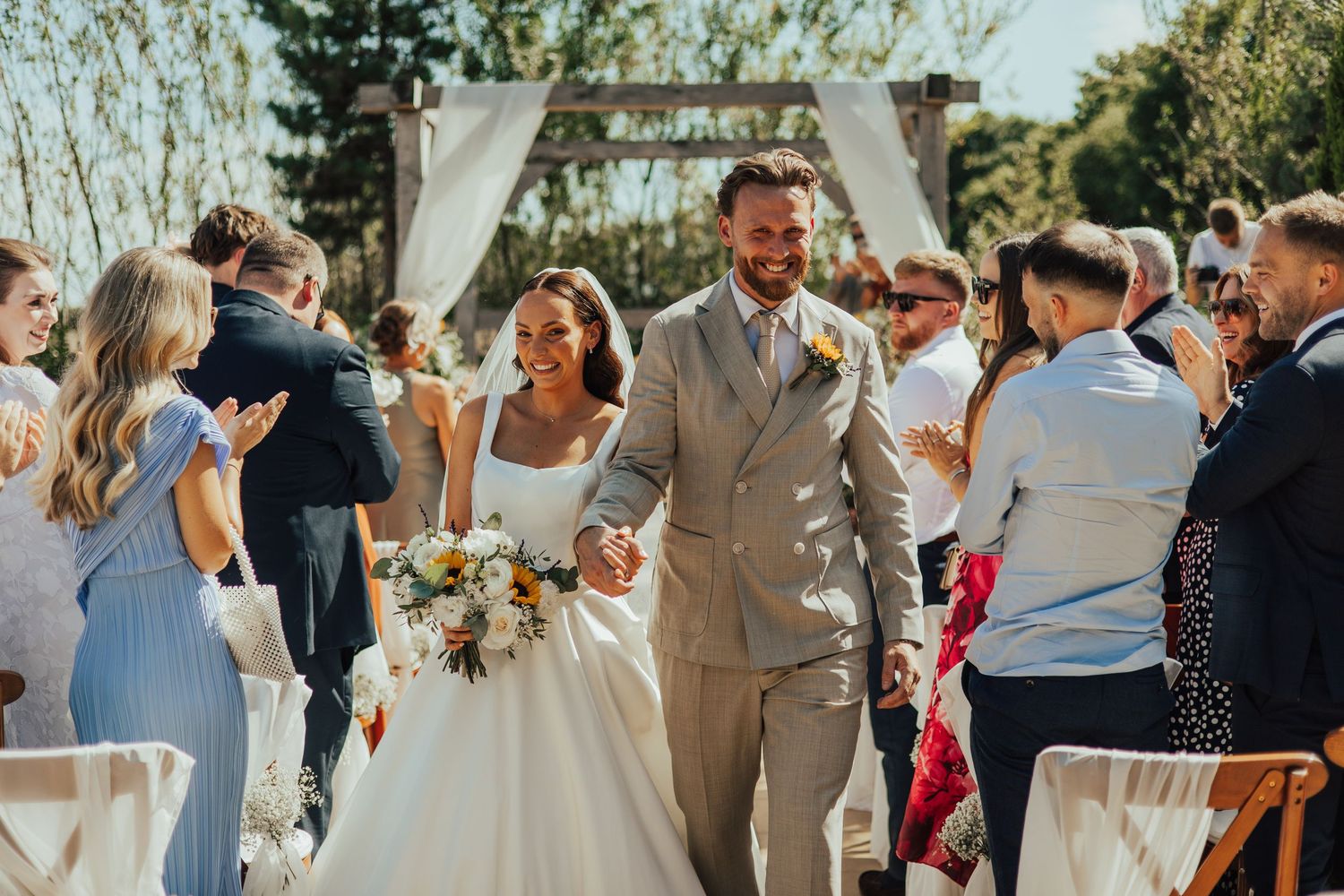 Guests mingle and celebrate at an outdoor wedding ceremony with natural sunlight filtering through the trees.