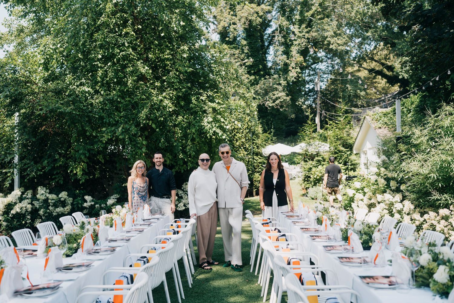 Garden party setup with white tables and chairs arranged beneath a canopy of leafy trees.