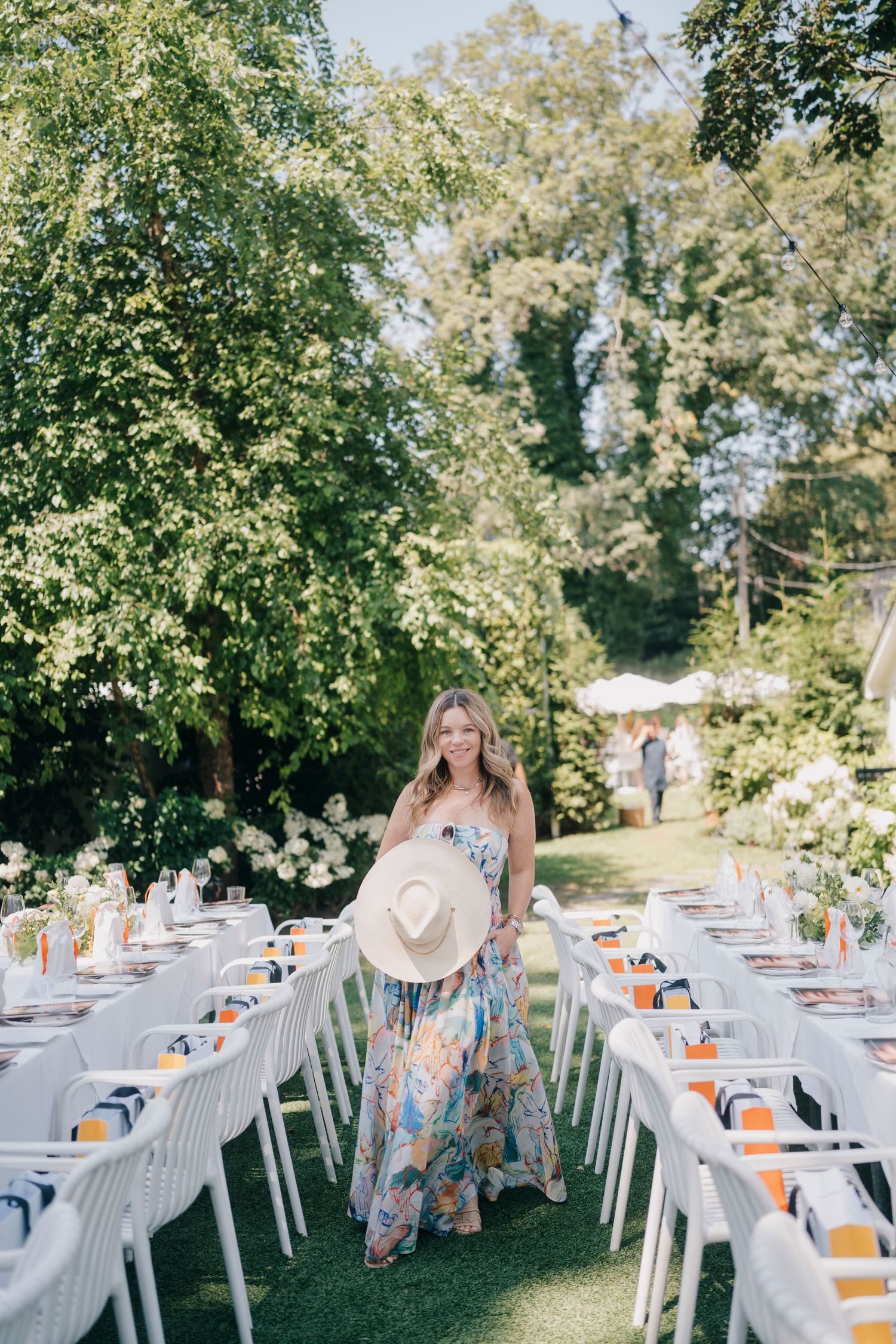 Elegant outdoor dining setup with white tables and orange accents under a tree canopy.