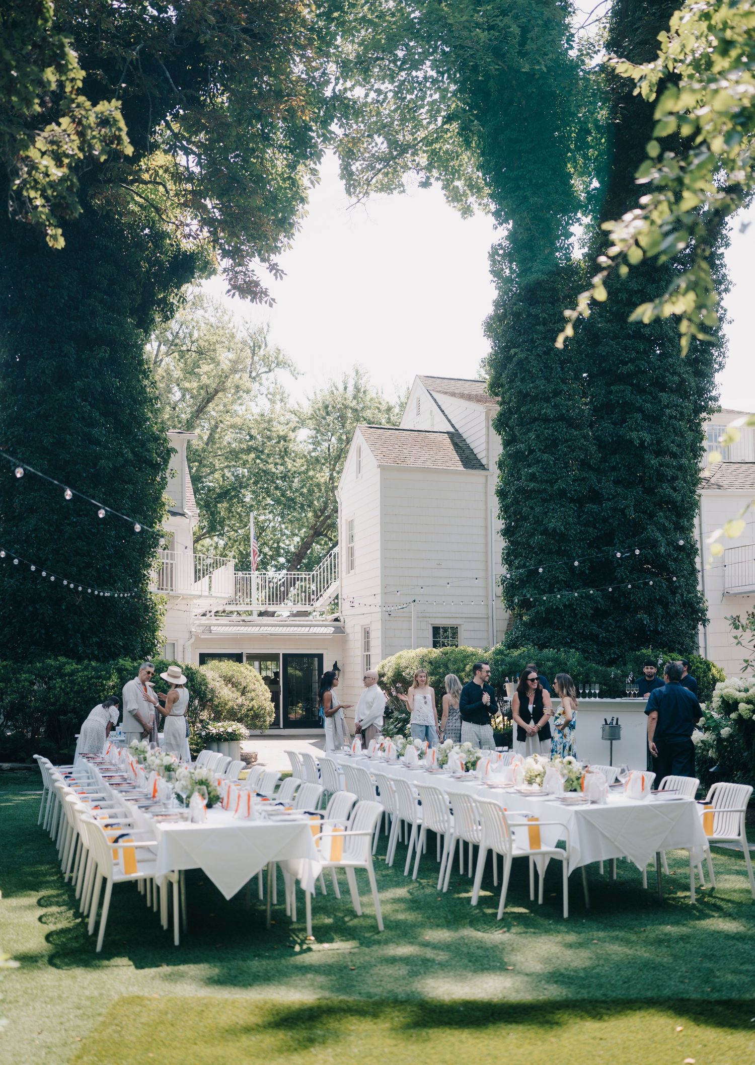 Garden reception area with white tables and chairs arranged beneath tall trees near white buildings.