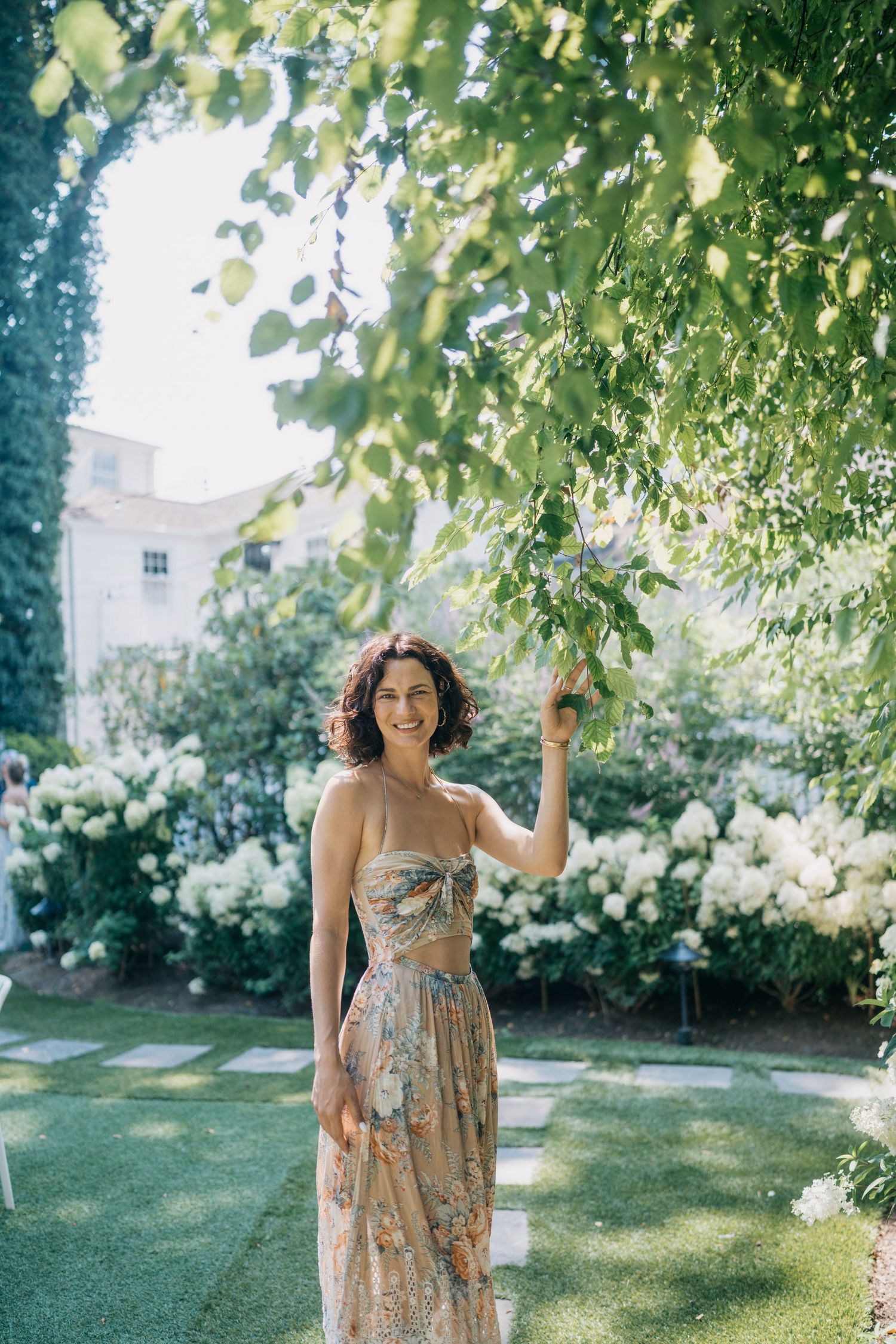 Woman in floral dress posing under green tree canopy at summer garden party.