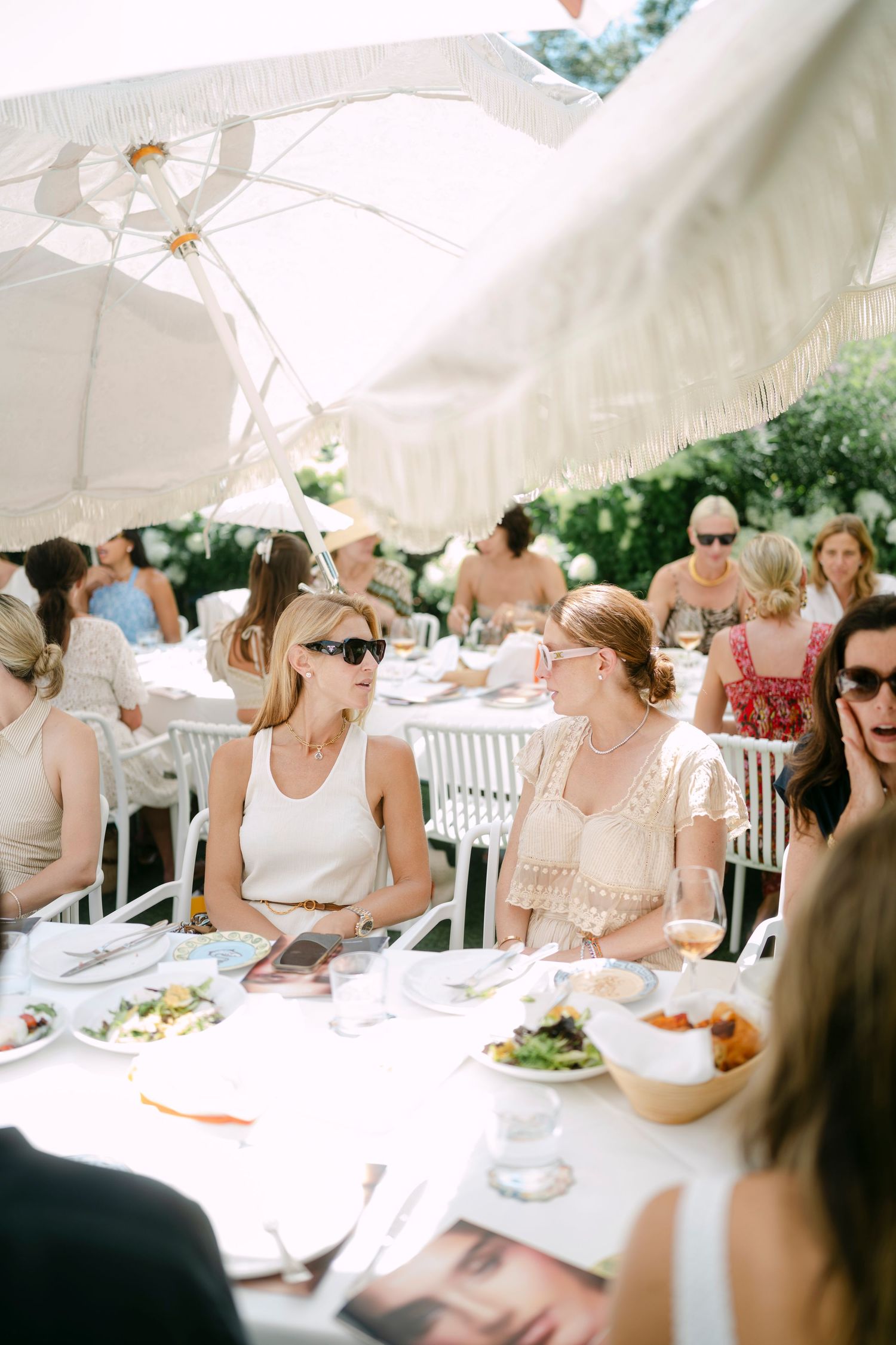Sun-drenched outdoor party scene with guests seated under white umbrellas at elegant tables.