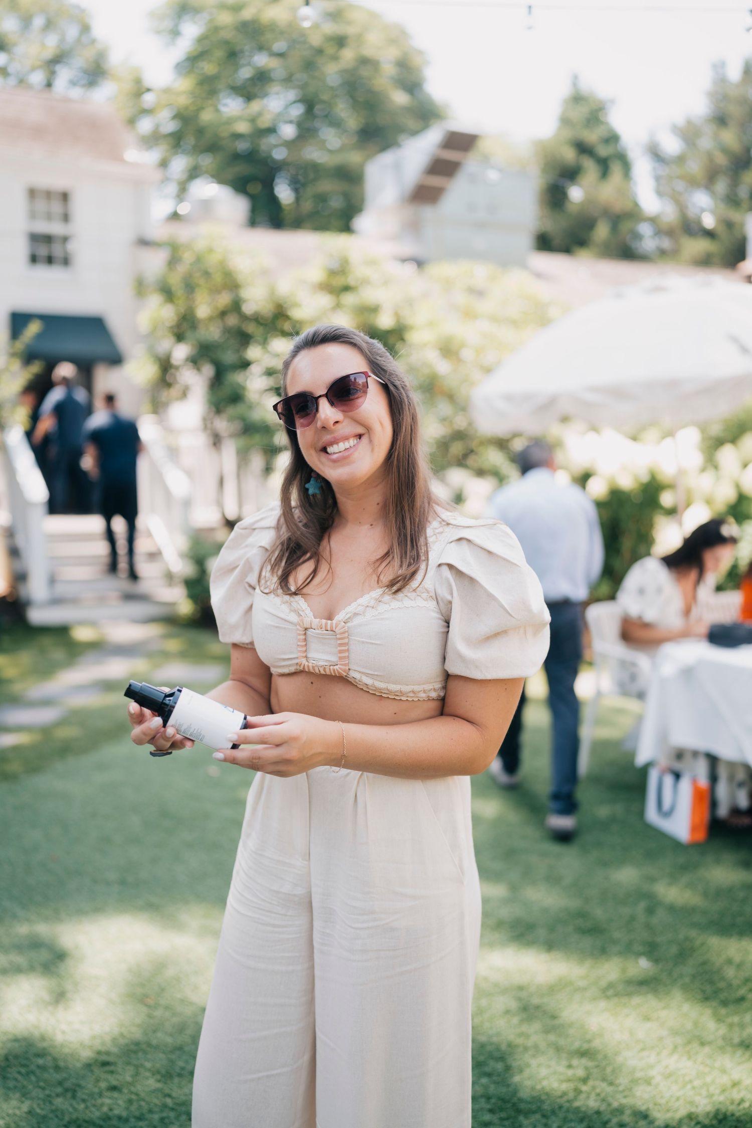 A guest in a cream-colored puff sleeve jumpsuit and sunglasses smiles at an outdoor summer party.