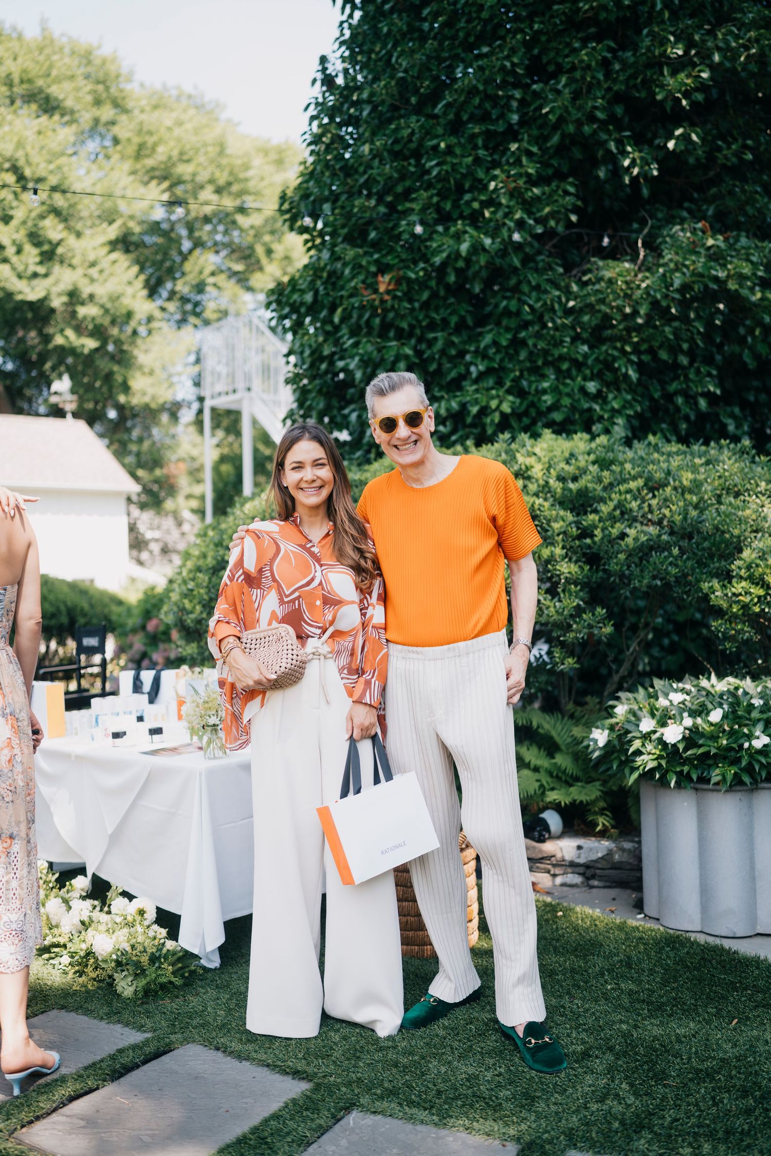 Two partygoers pose together, one wearing an orange printed top and one in bright orange, at an outdoor event.