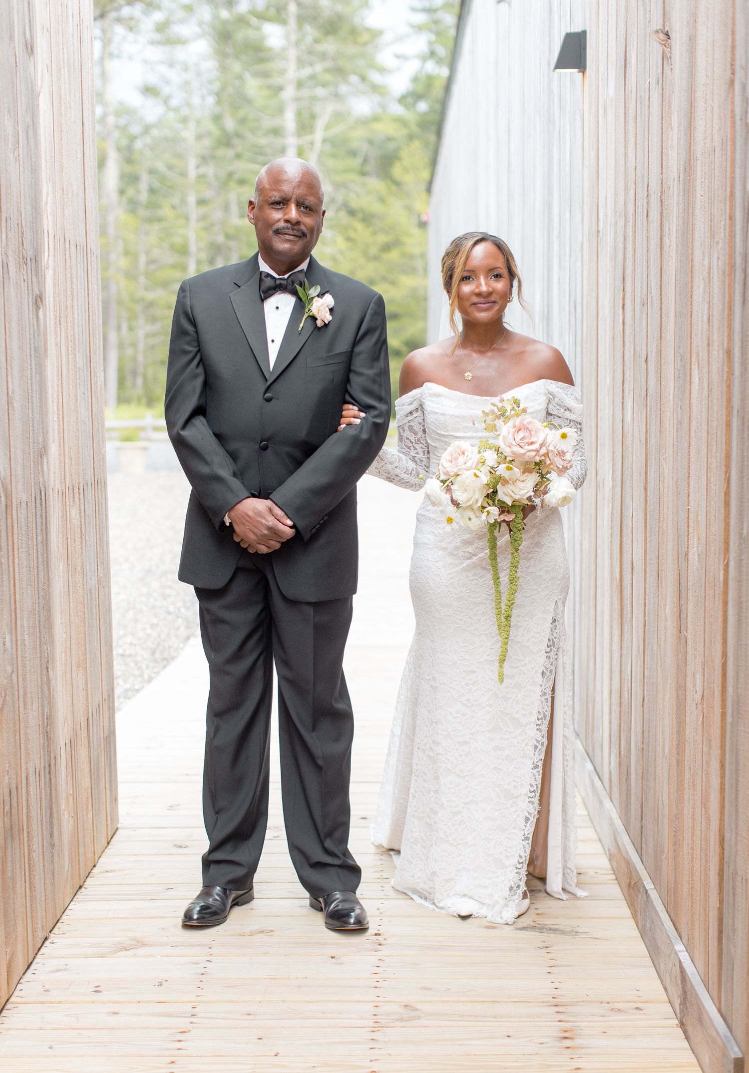 A wedding ceremony takes place under a decorated floral arch on a wooden deck surrounded by guests and forest scenery.