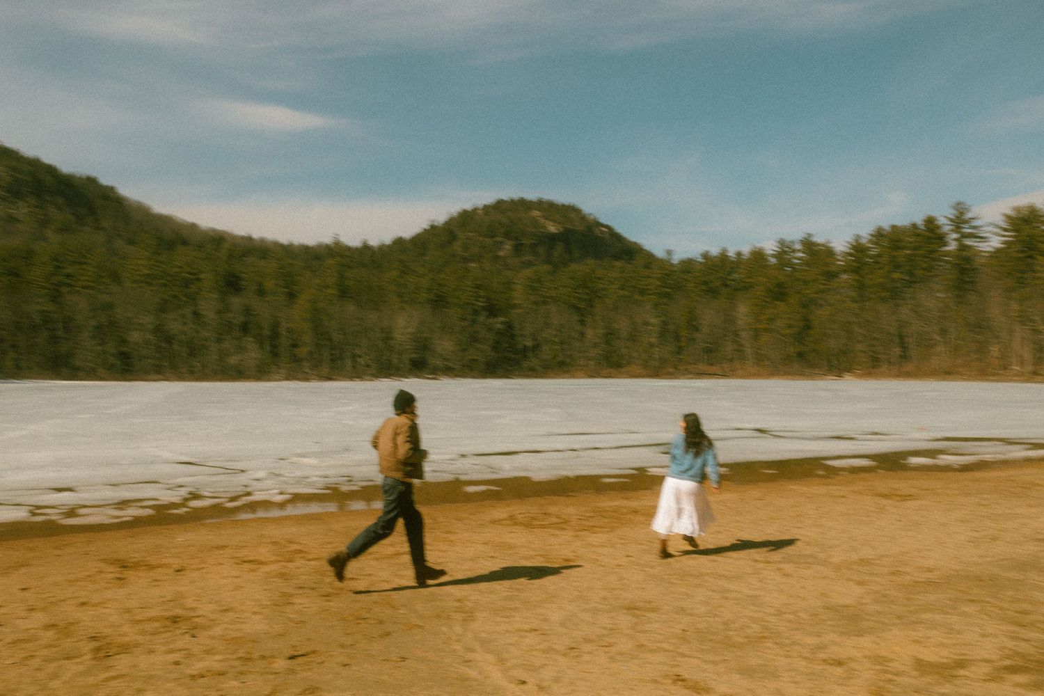 Two people walking along a snowy beach with mountains in the background.