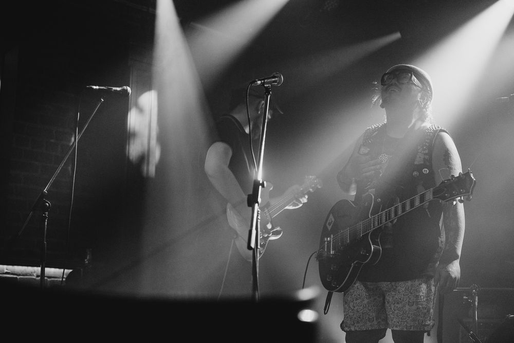 Black and white concert photo showing silhouetted band members performing under dramatic spotlights on stage.
