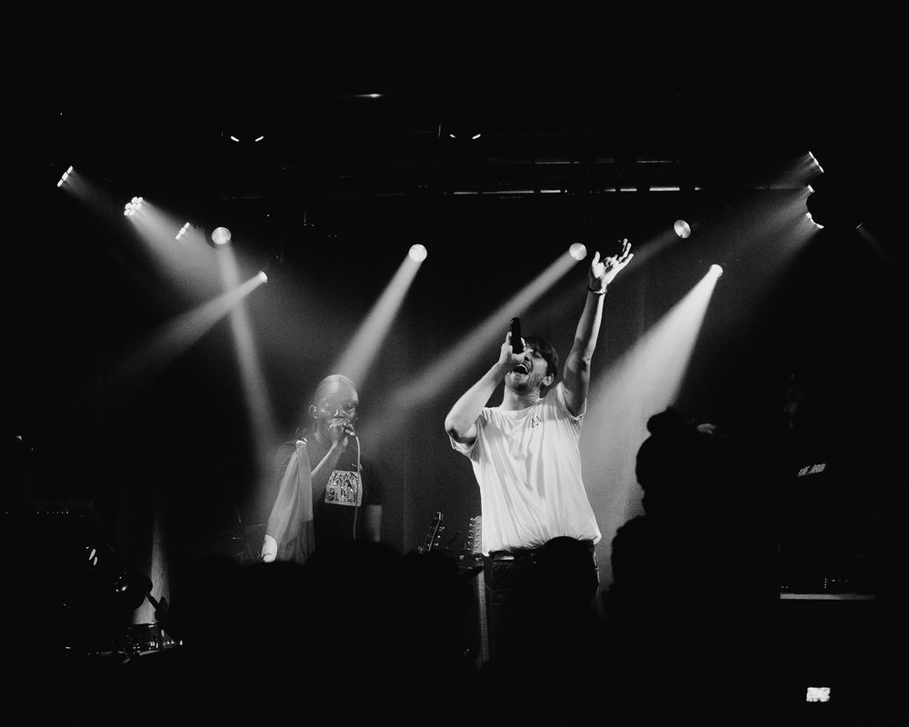Dark concert stage illuminated by dramatic spotlights during an energetic hip-hop performance.
