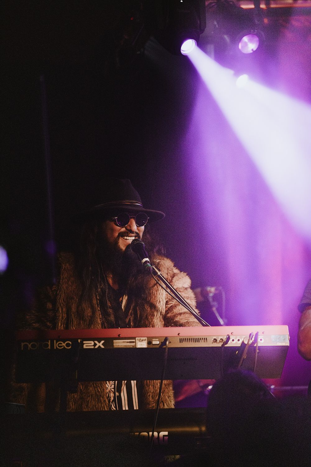 Musician performs on keyboard in atmospheric purple stage lighting during live concert performance.
