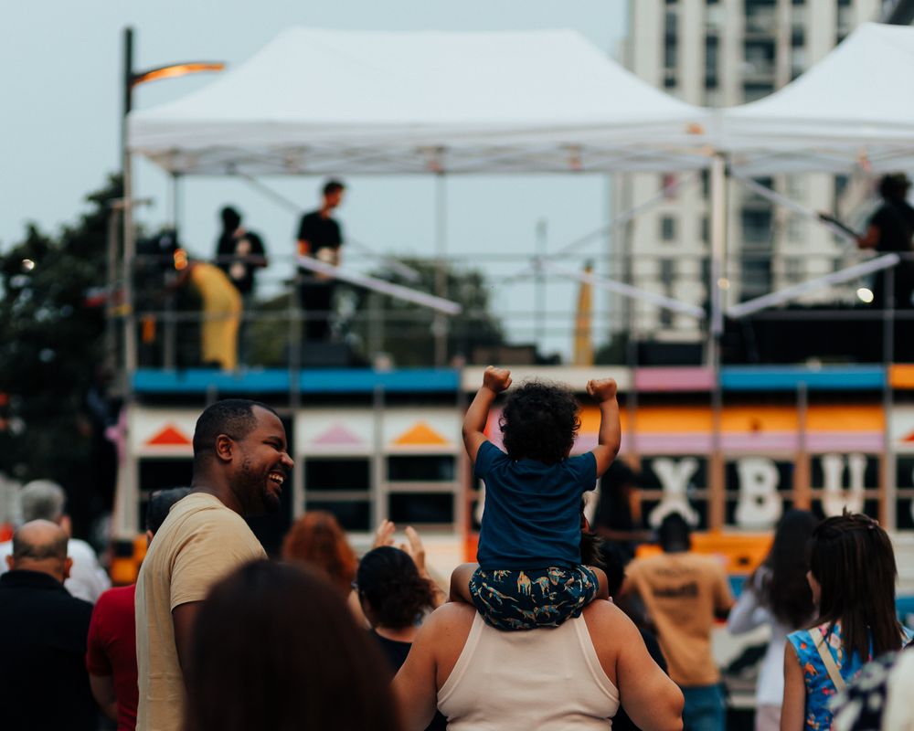 Group of people watching from a crowd at an outdoor event with buildings in the background.