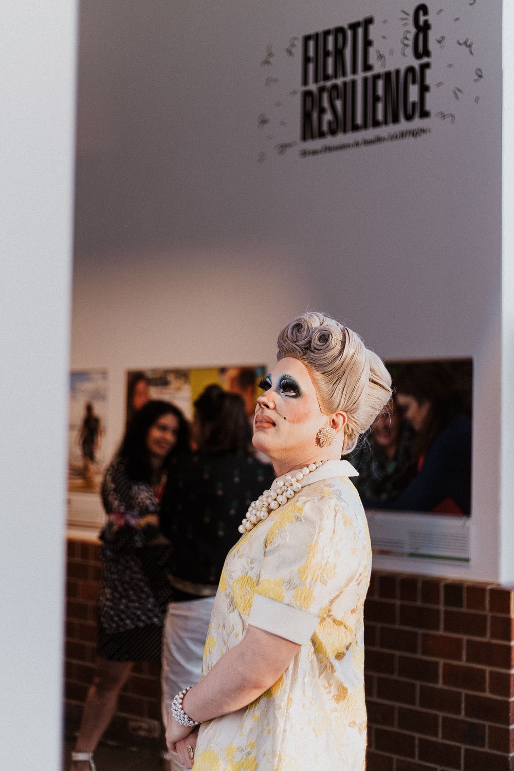 Person in light-colored outfit and accessories leans against white gallery wall with text signage.