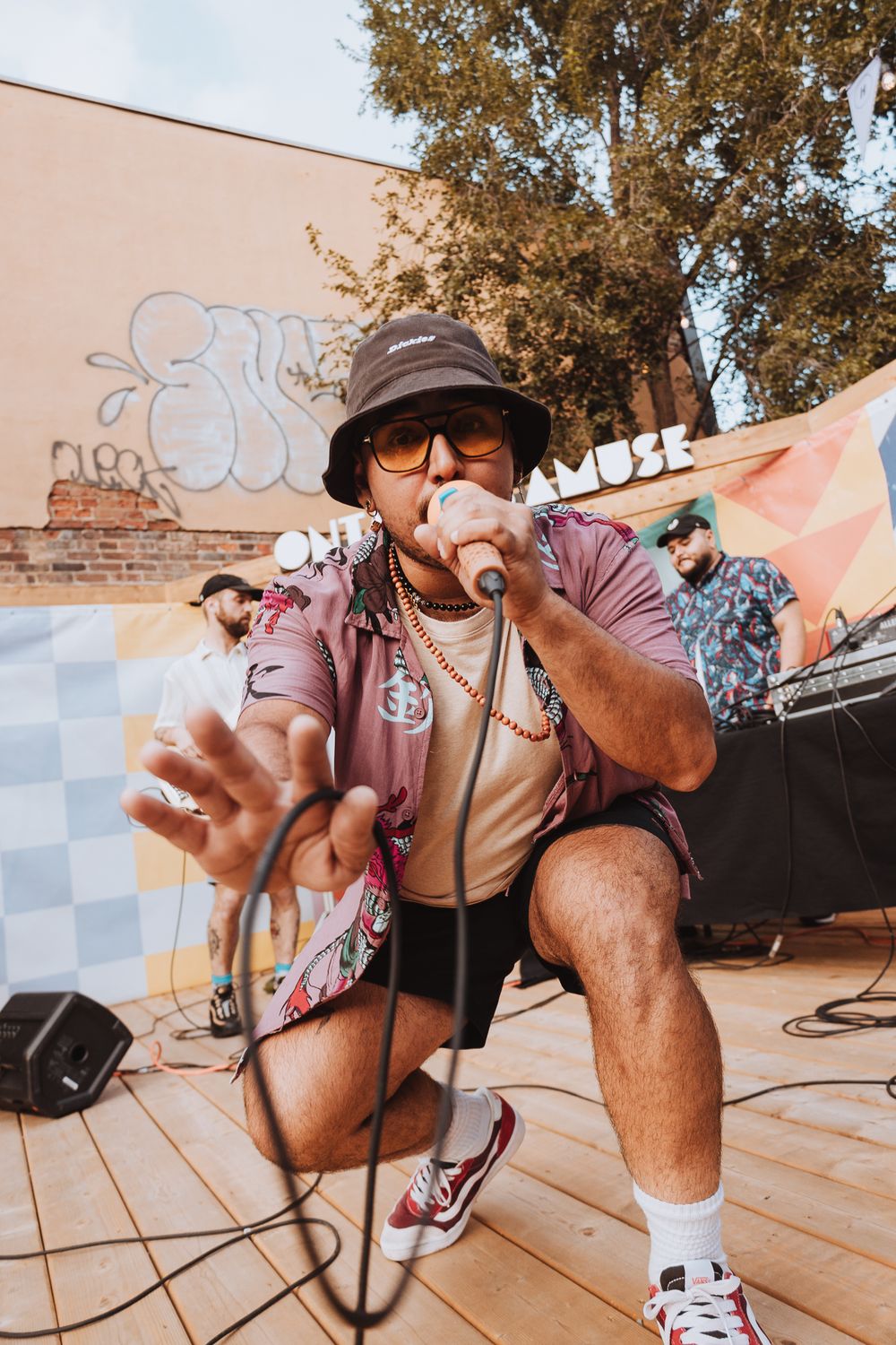 A performer in casual attire and bucket hat crouches while using audio equipment outdoors near graffiti walls.