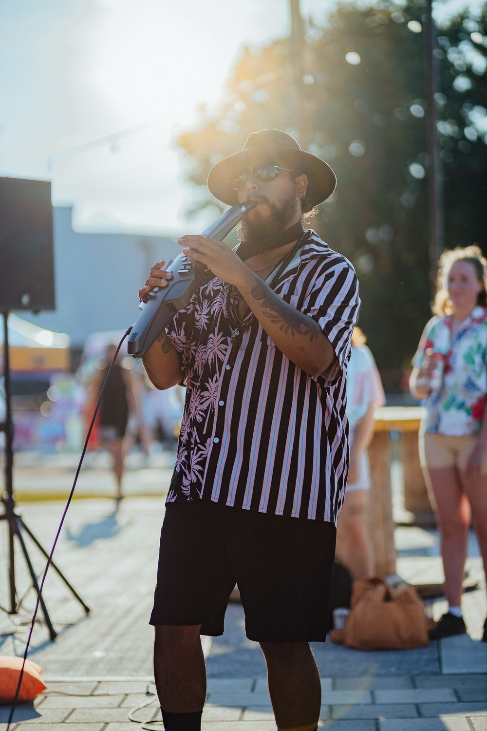 Musician in striped shirt and hat performs outdoors at a summer festival in backlit sunlight.