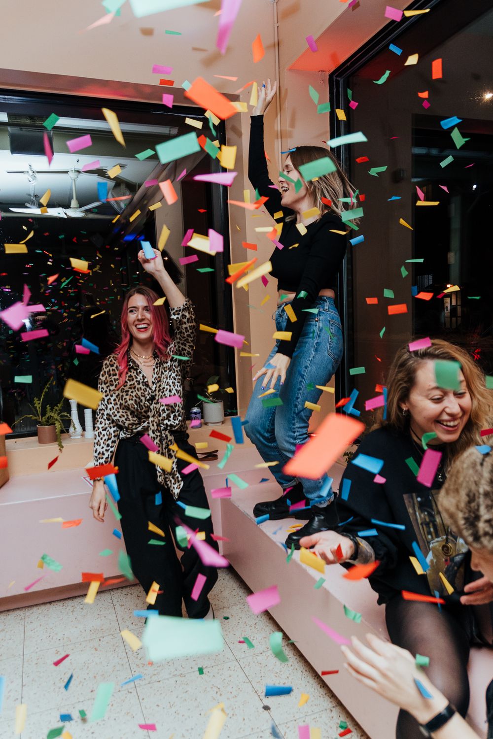 People celebrate at a party with colorful confetti falling and dancing in a dimly lit room with red lighting.